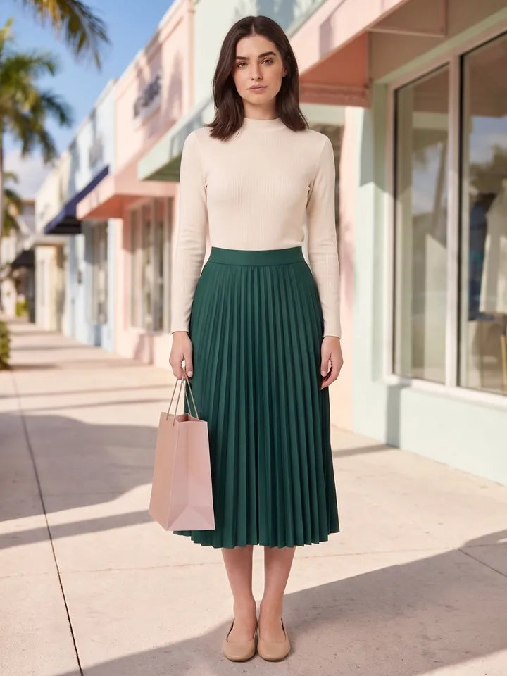 A woman in a green pleated midi skirt and a cream knit top, holding a pink shopping bag on a sunny street