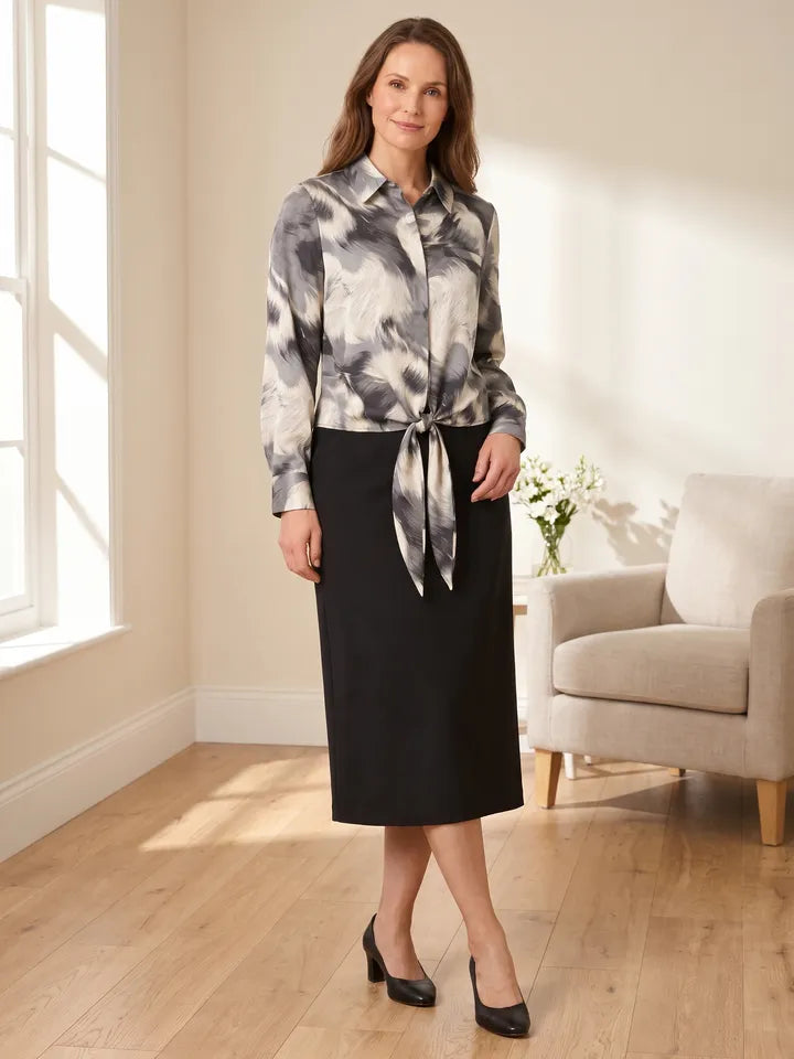 A woman in a grey and cream print tie-front blouse and a black midi skirt in a bright room