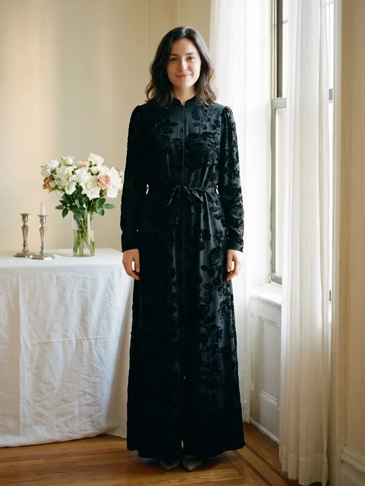 A woman in the Modest Robe Front Zipper Black Velvet Floral by a window, beside a table with white flowers and candlesticks